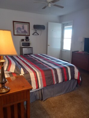Un dormitorio con cama, escritorio, lámpara, microondas, perchero y una puerta.