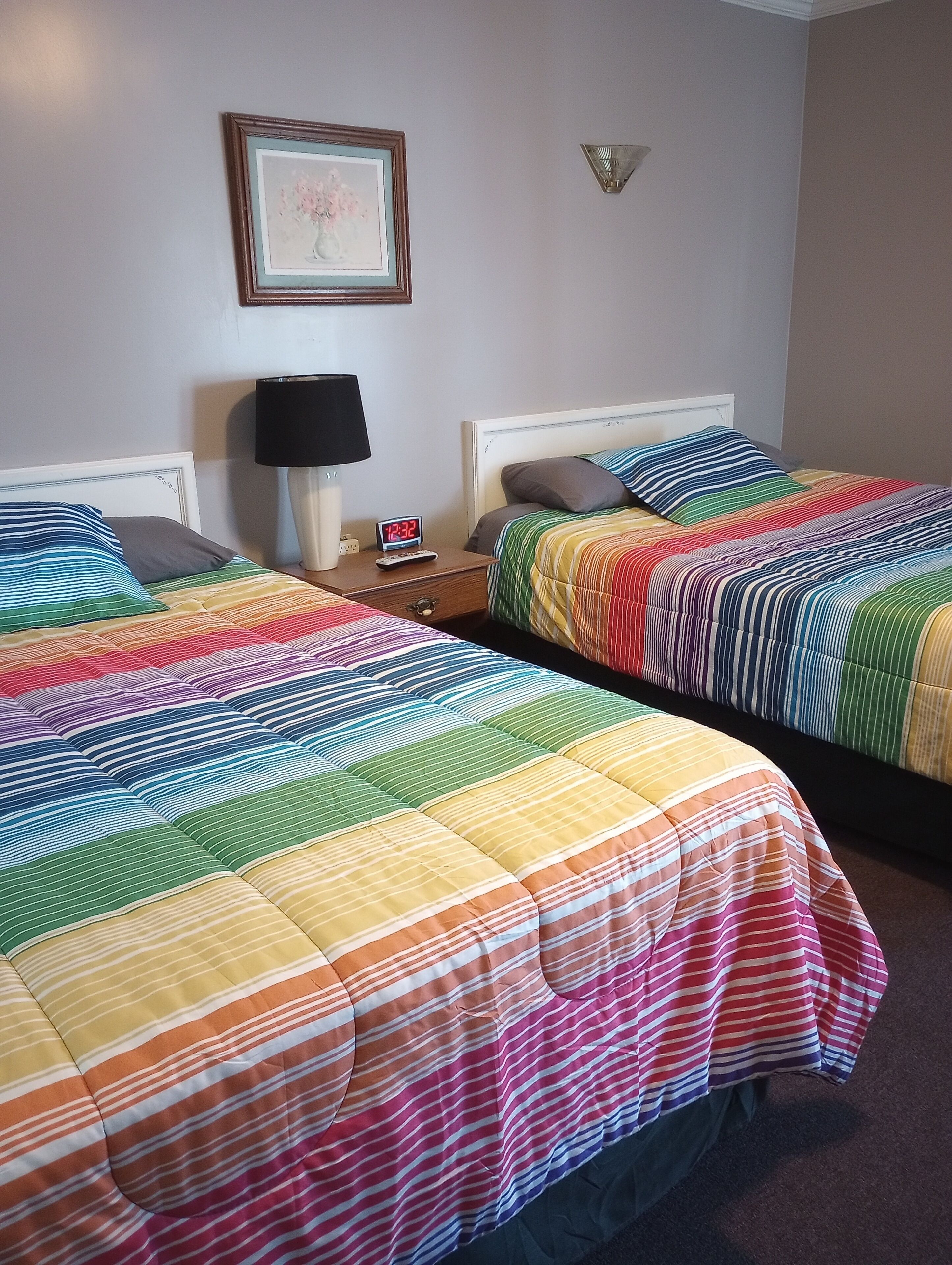 Two beds with colorful striped bedding, a nightstand with a lamp and a digital clock, and a framed picture on the wall.