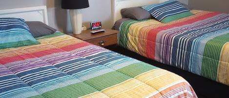 Dos camas con ropa de cama a rayas coloridas, una mesita de noche con una lámpara y un reloj digital, y un cuadro en la pared.