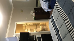 A compact hotel room with a bed, a small desk, a chair, a wardrobe, and a bathroom visible through an open door.