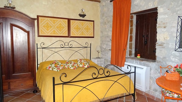 Un dormitorio con cama, armario, radiador, ventana con cortinas y una mesita con una cesta.