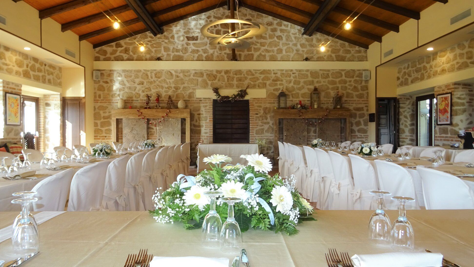 Un salón de banquetes con paredes de piedra, vigas de madera y chimenea. Las mesas están dispuestas con manteles blancos y cristalería.