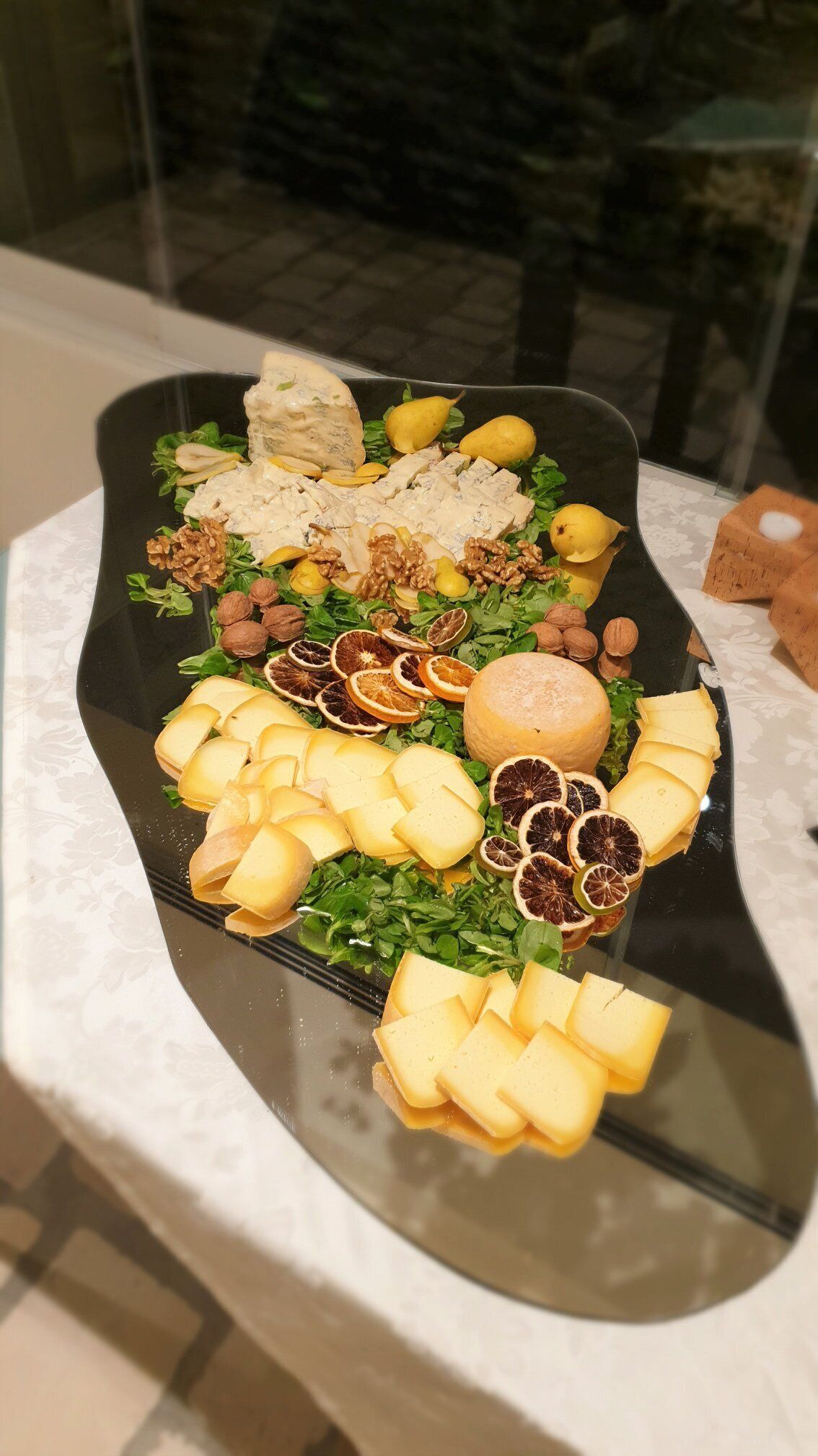 Una bandeja con quesos variados, frutos secos y frutas deshidratadas sobre una mesa de vidrio.
