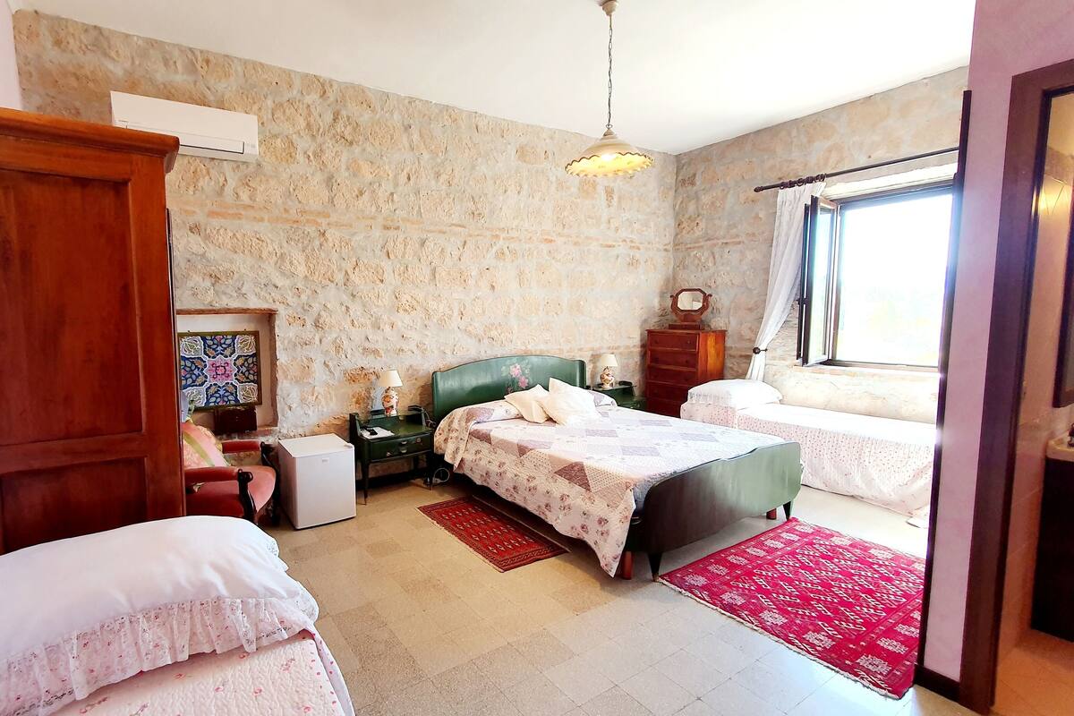 A bedroom with a stone wall, a wooden wardrobe, a bed with a floral bedspread, a nightstand with a lamp, and a window with curtains.