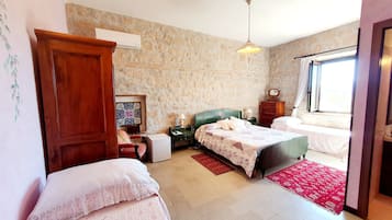 A bedroom with a stone wall, a wooden wardrobe, a bed with a floral bedspread, a nightstand with a lamp, and a window with curtains.