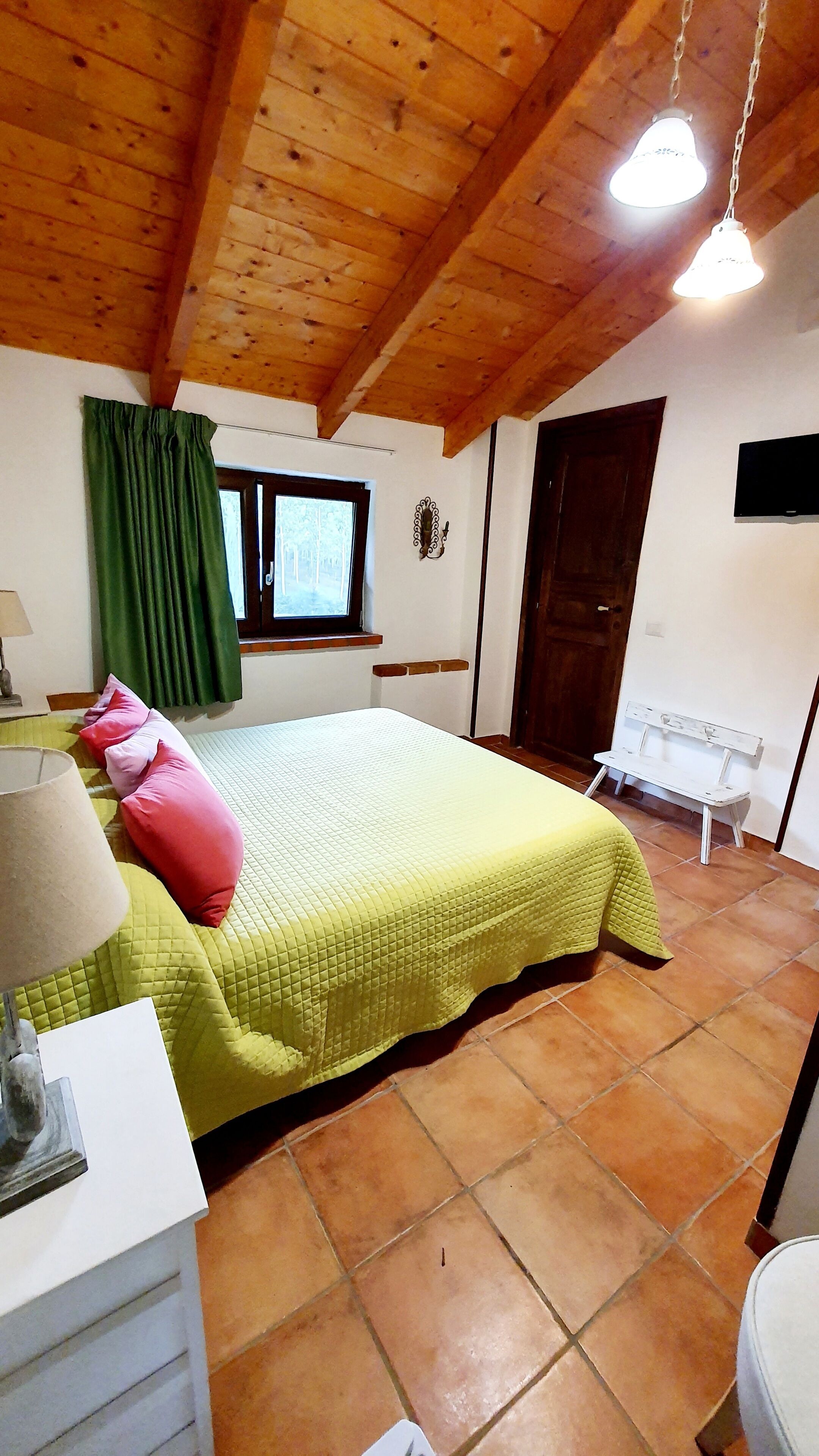 Ein Schlafzimmer mit Bett, grünen Vorhängen, Holzboden, einem Fenster, einer Bank und einer Tür.
