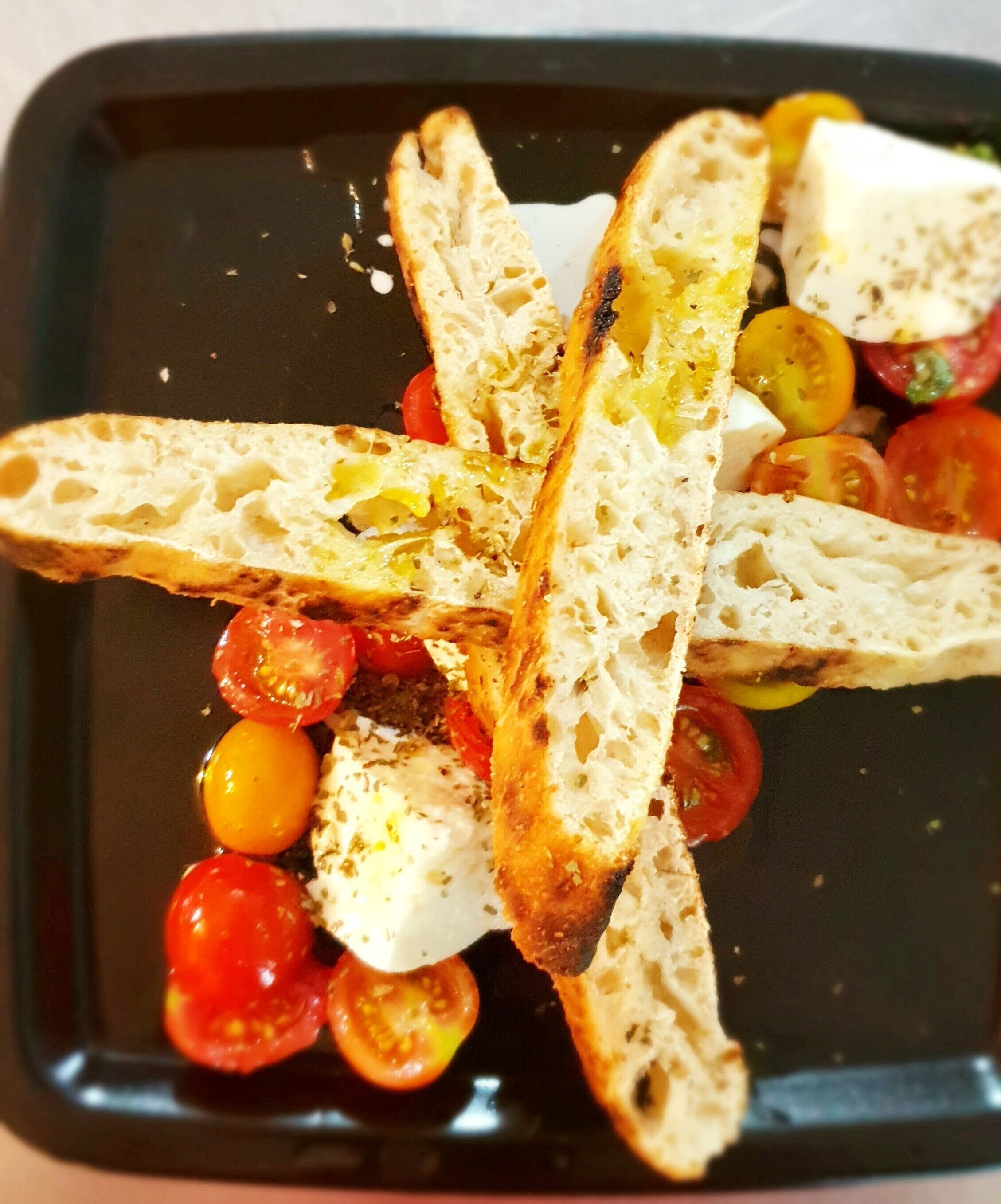 Un plato negro con pan a la parrilla, tomates cherry y una cobertura cremosa blanca.