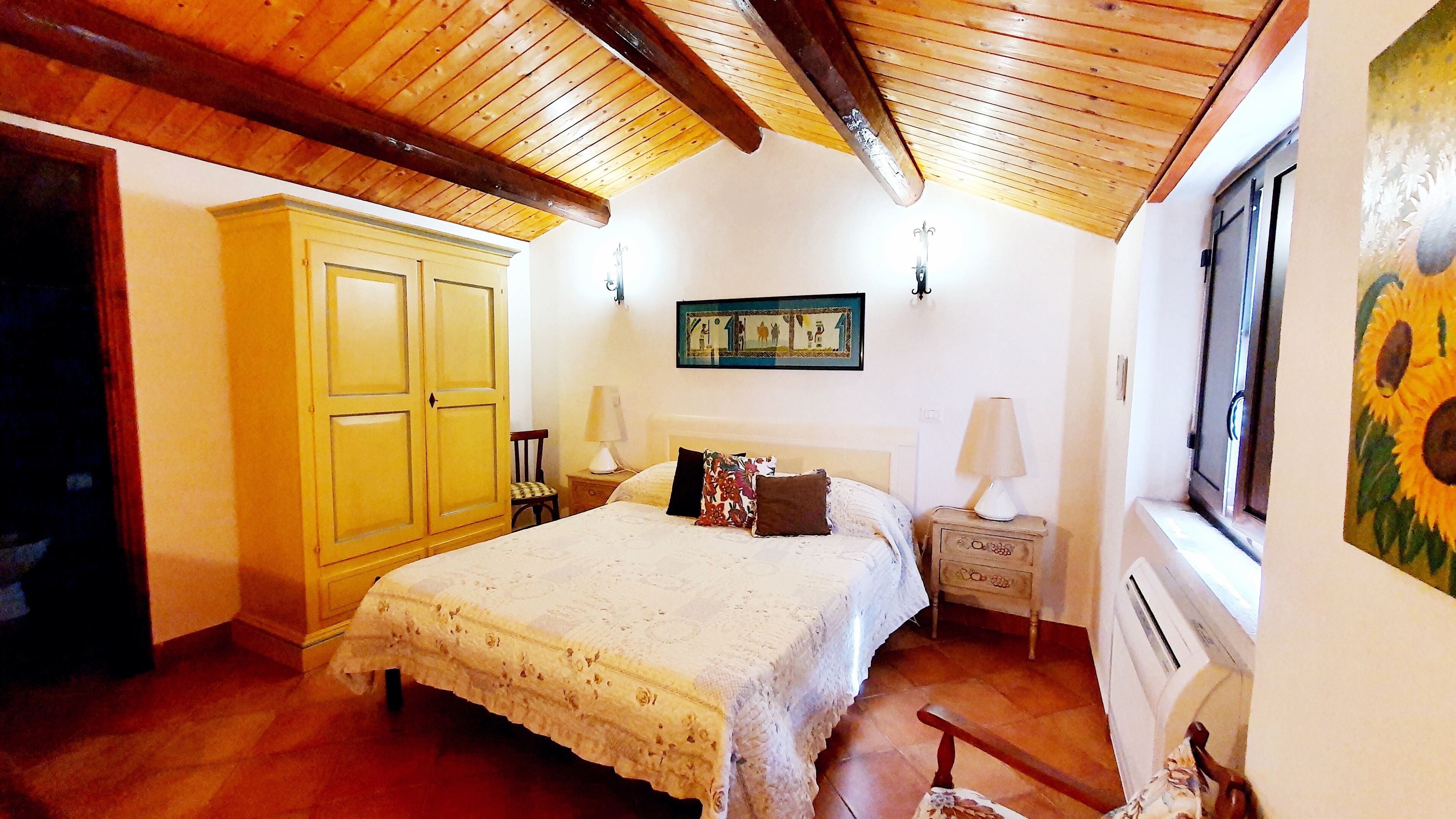 A bedroom with a wooden ceiling, a bed with a white bedspread, two bedside lamps, a chair, and a painting of sunflowers.