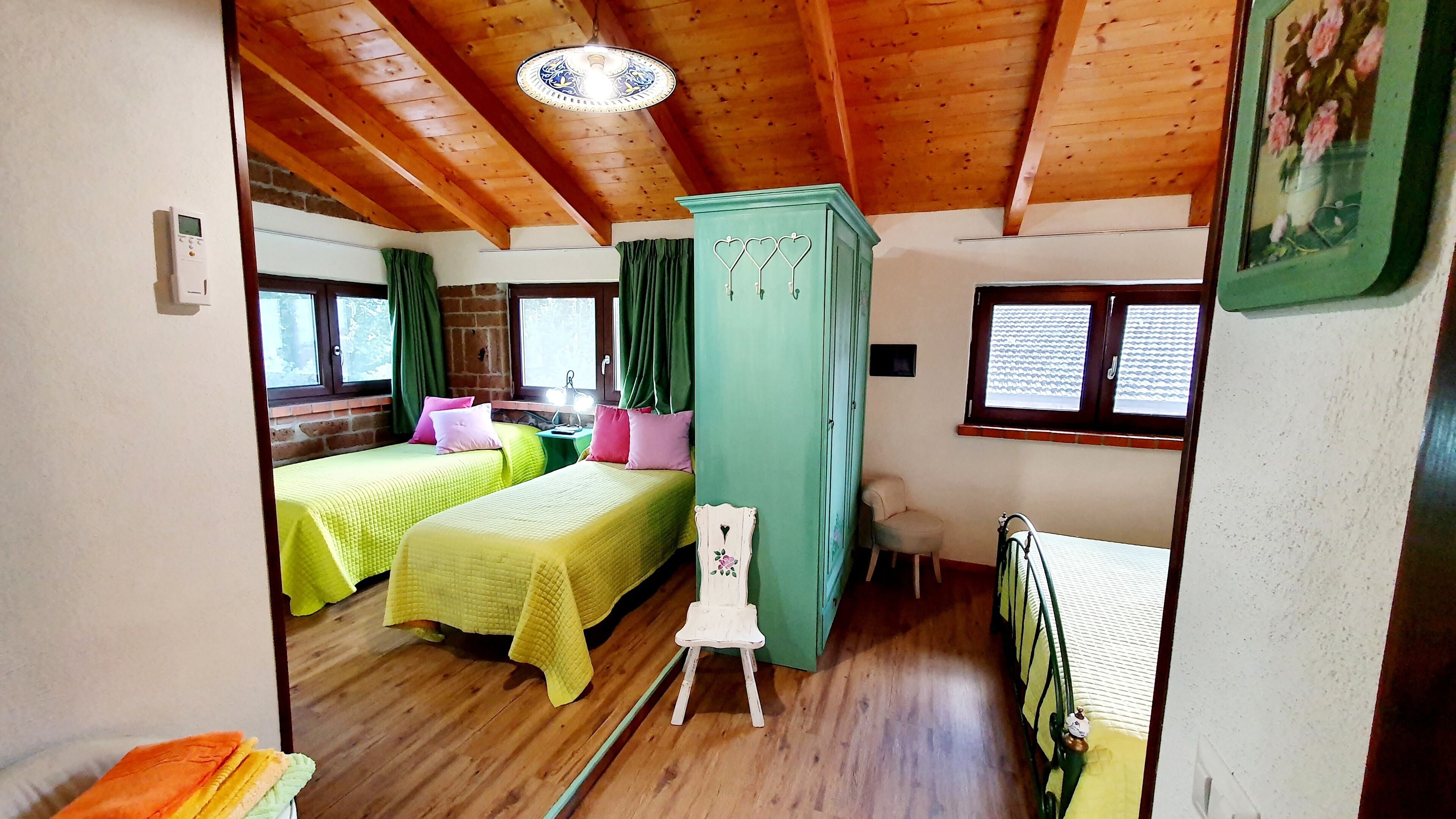 Quarto com duas camas, um guarda-roupa verde, uma cadeira branca pequena e uma janela com persianas.