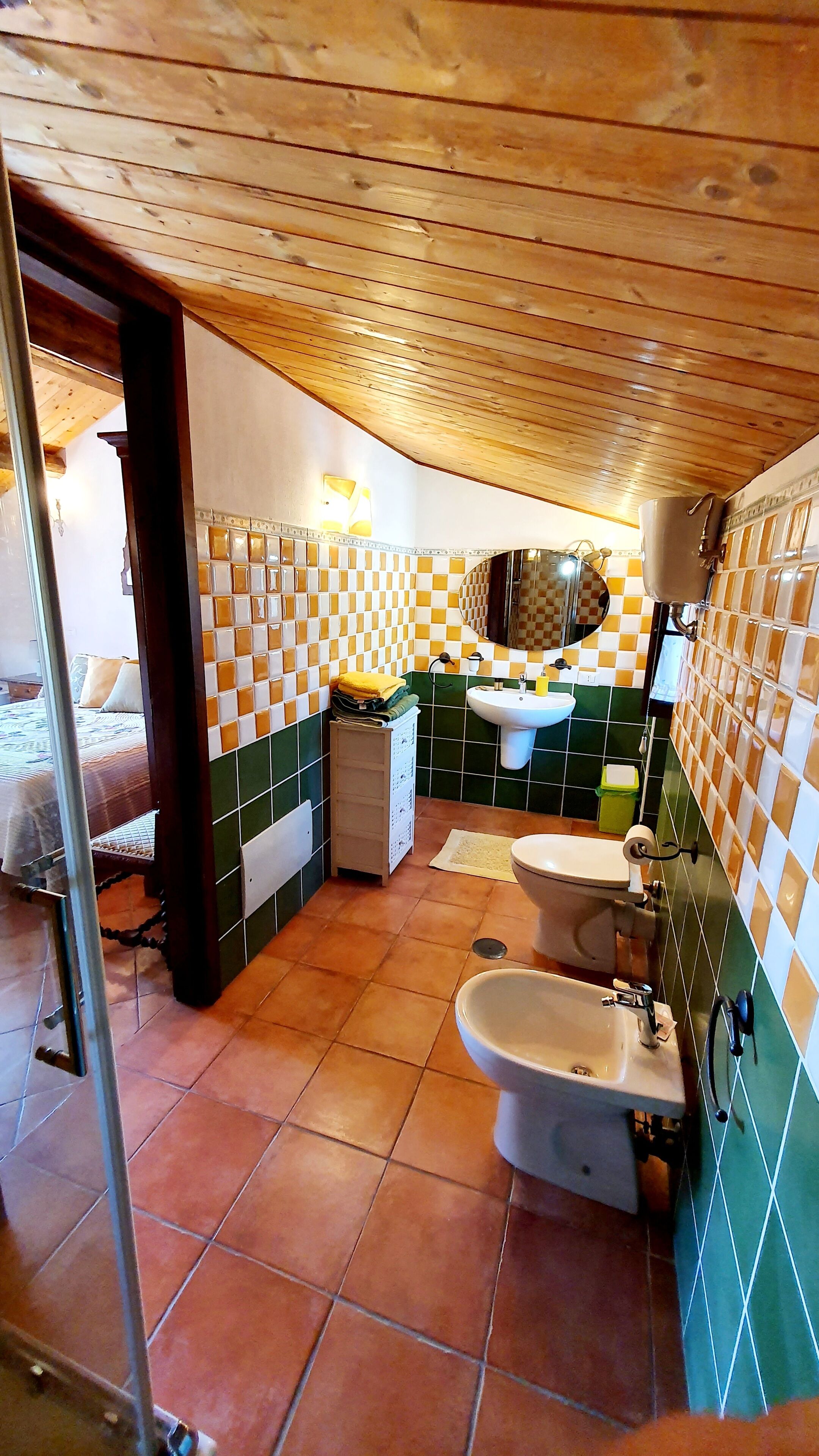 Un baño con lavamanos, inodoro y bidé, con azulejos verdes y blancos, techo de madera y una cama visible al fondo.