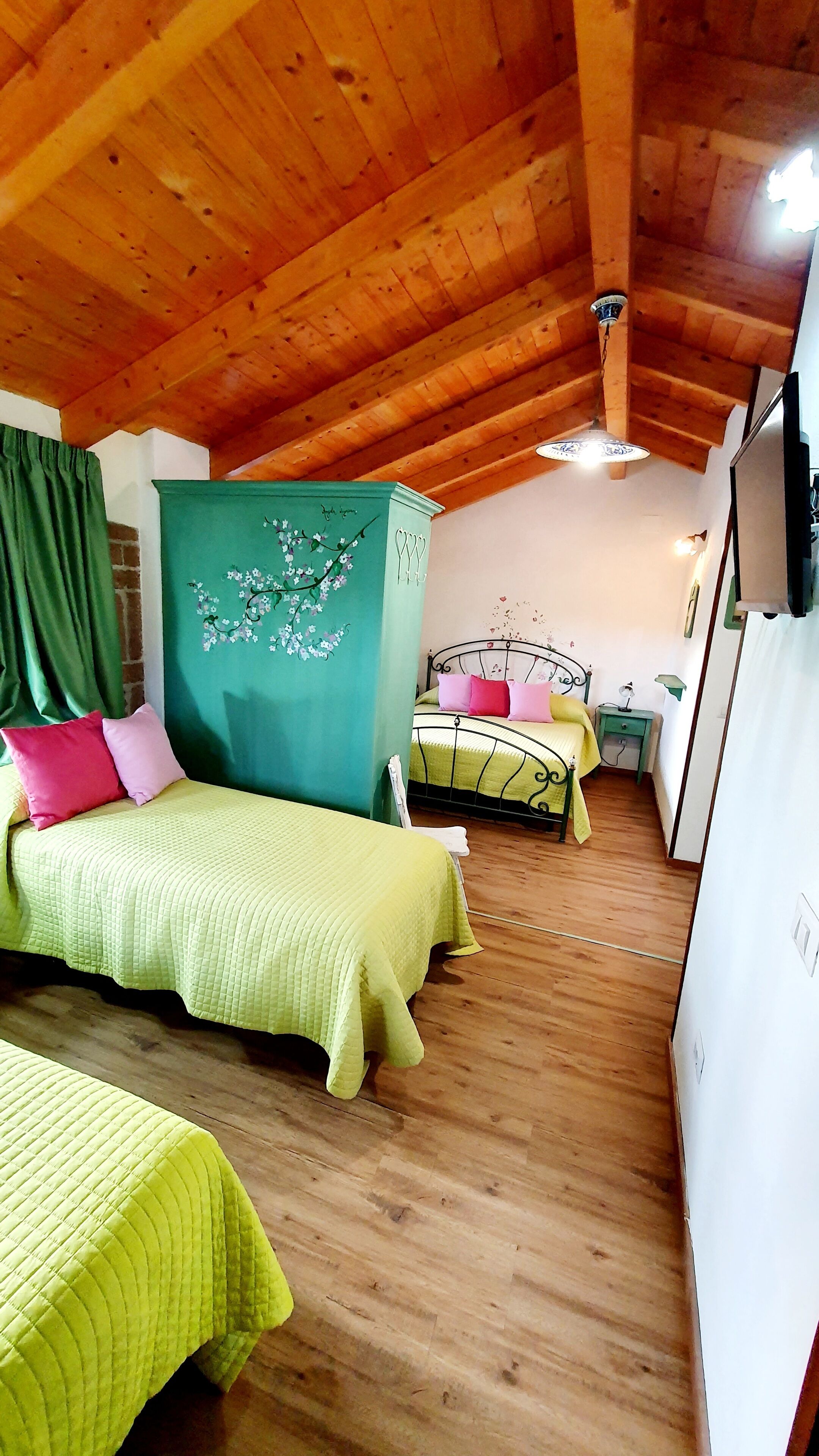 A cozy bedroom with wooden beams, two beds with green and yellow bedding, a green headboard, and a small bedside table with a lamp.