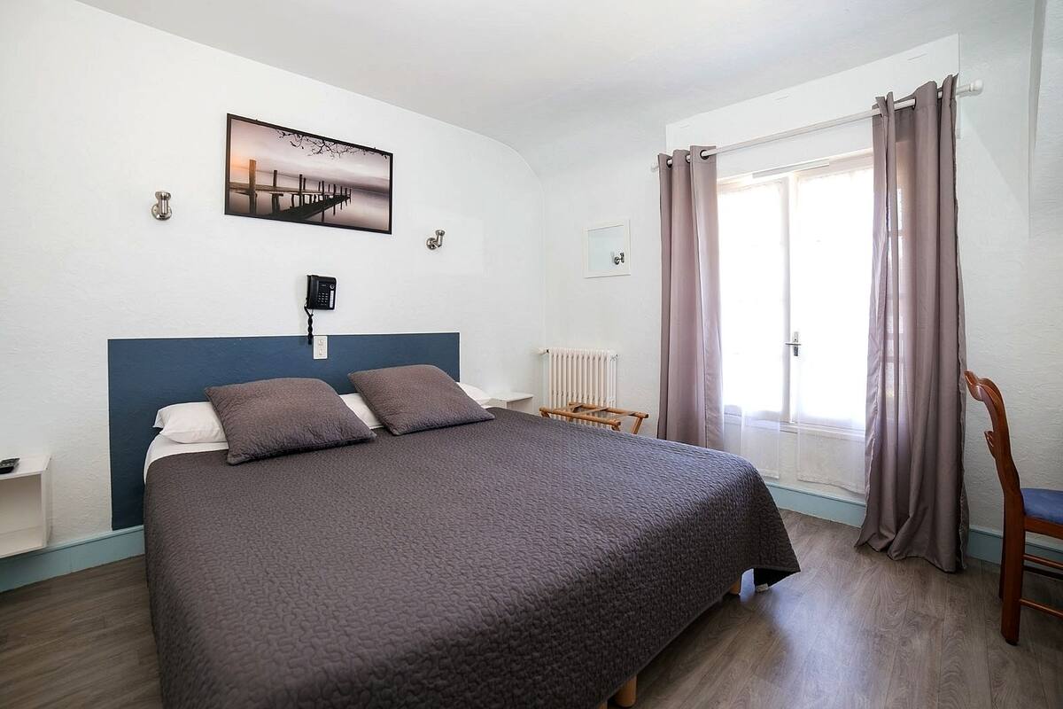 A bedroom with a bed, a bedside table, a phone, a radiator, a window with curtains, and a chair.