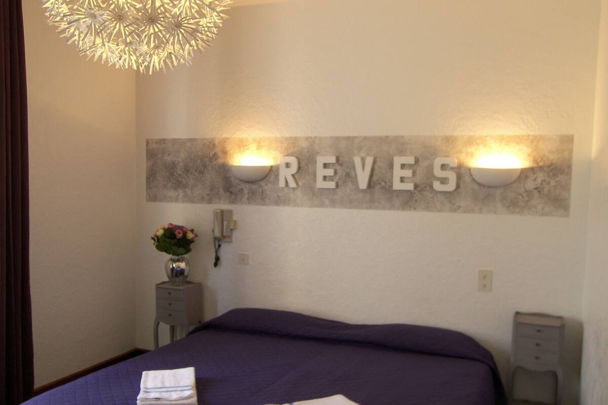 A hotel room with a bed, two wall-mounted lights, a decorative wall piece spelling "REVES", a vase with flowers, and a telephone on a nightstand.