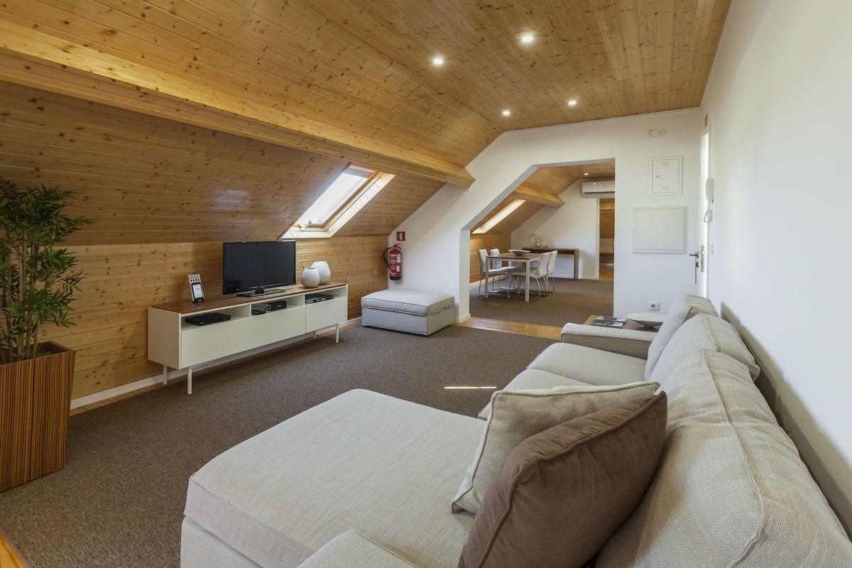 A modern loft-style living room with a high ceiling and skylights, a white sofa, a TV on a white console, and a dining area in the background.