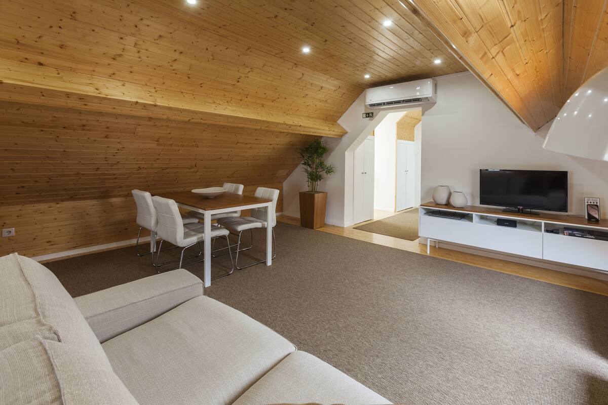 A modern loft-style room with a wooden ceiling, a dining area with a white table and chairs, a sofa, a TV on a white entertainment unit, and a potted plant.