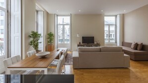 A modern living room with a wooden dining table, white chairs, a beige sofa, and a TV. - BO - Fernandes Tomás (Porto)