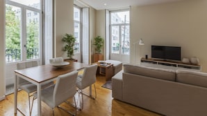 A modern living room with a wooden dining table, white chairs, a beige sofa, a TV, and large windows with a view of buildings and greenery. - BO - Fernandes Tomás (Porto)