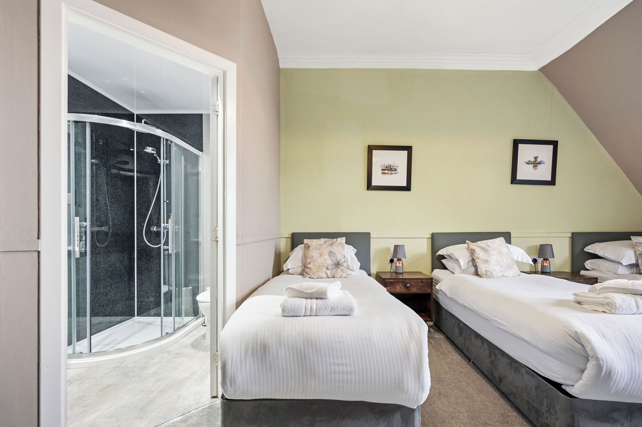 Une chambre d’hôtel dotée d’une douche en verre, de deux lits avec du linge de lit blanc, d’une table de chevet avec une lampe et de deux tableaux encadrés au mur.