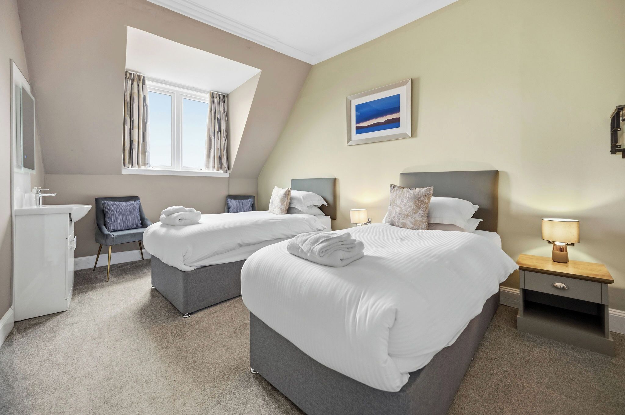 A hotel room with two beds, a desk, a chair, a window, and a painting on the wall.