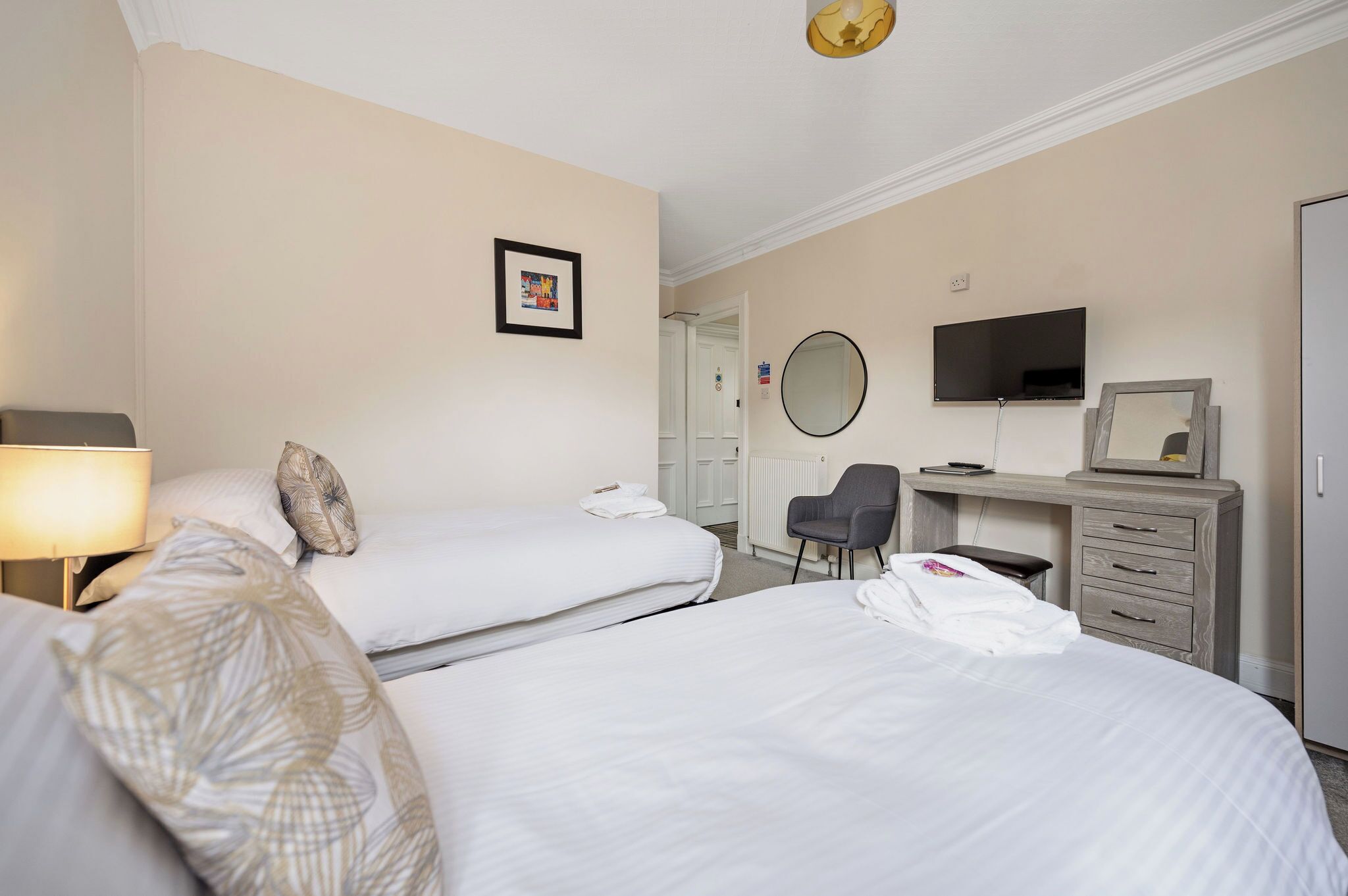A hotel room with two beds, a desk with a chair, a TV, a mirror, and a lamp.