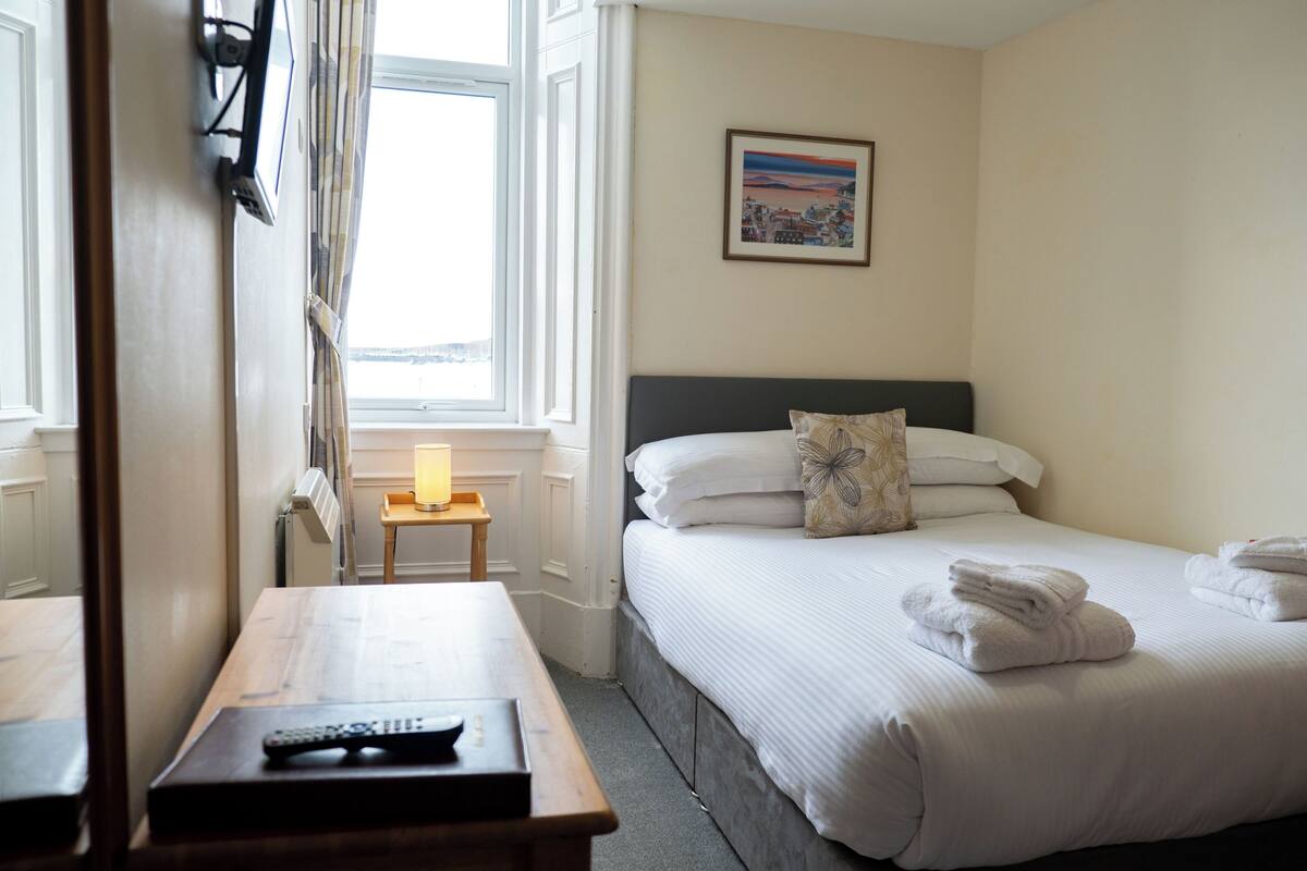 A hotel room with a bed, a bedside table with a lamp, a remote control, and a window with curtains.