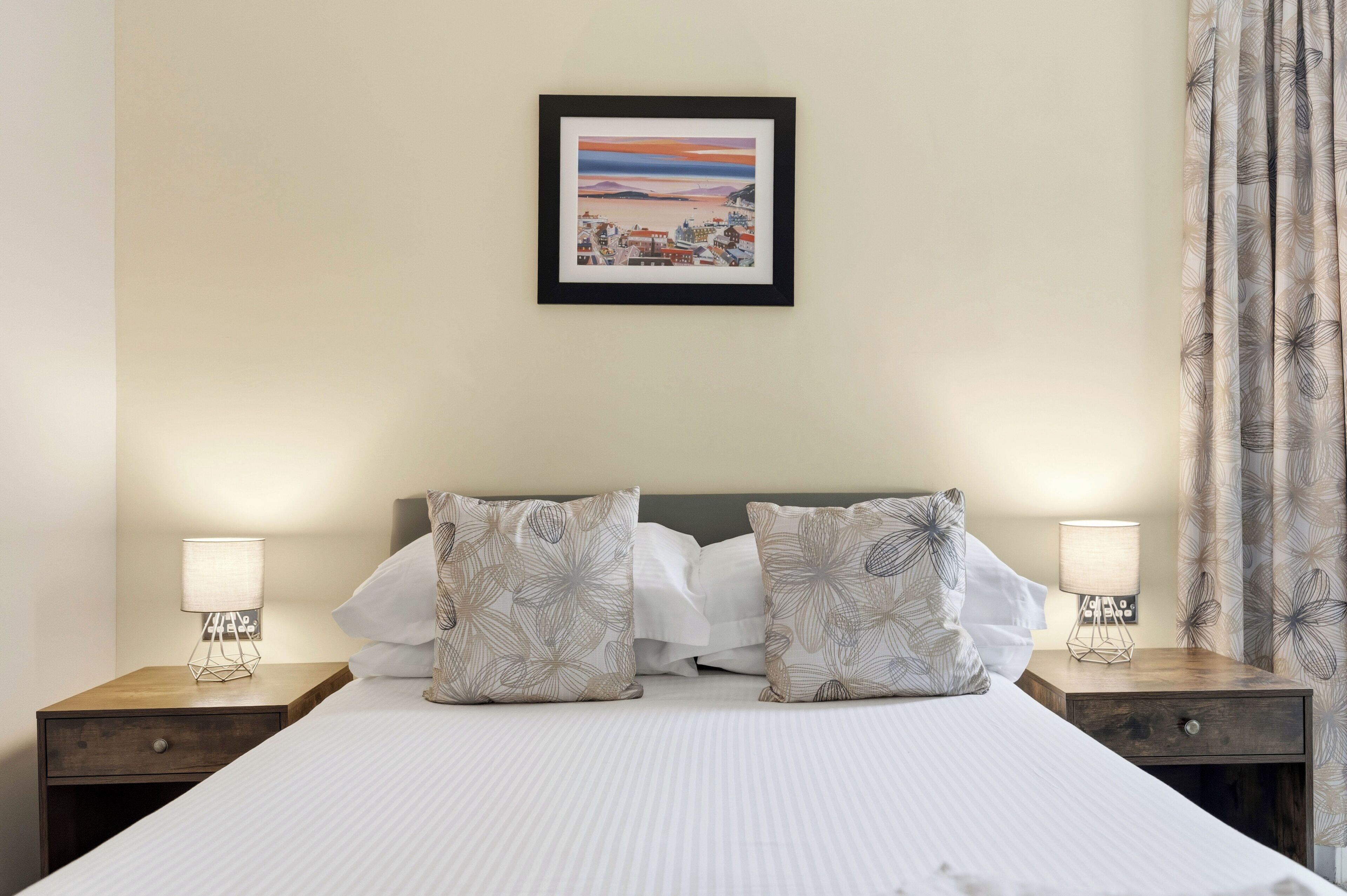 A neatly made bed with white linens, two matching pillows, a framed picture on the wall, and a nightstand with a lamp and a drawer.