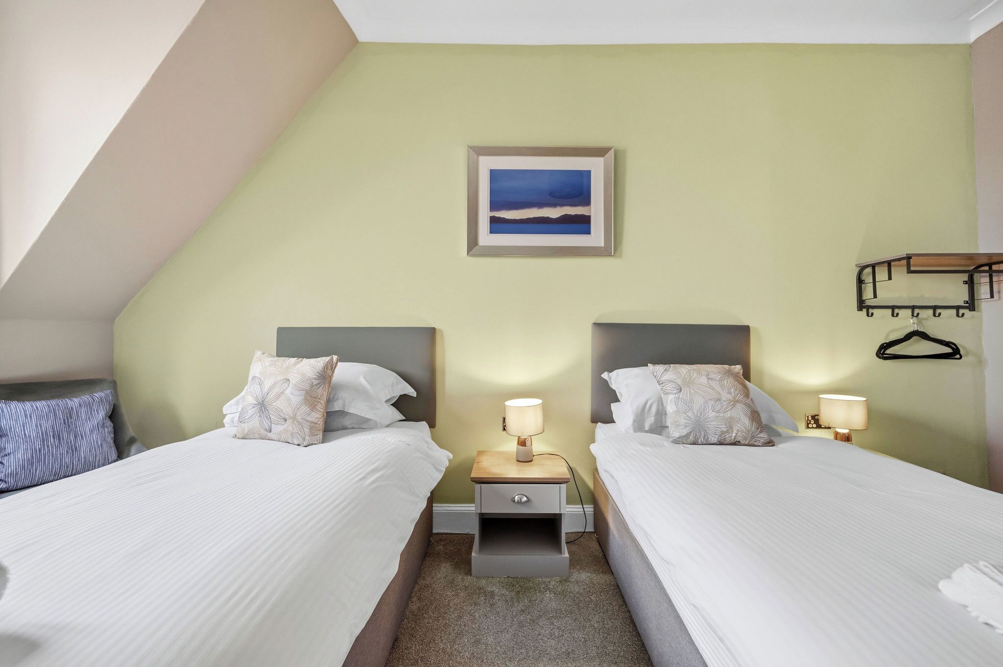 Une chambre double avec deux lits, une table de chevet avec une lampe, un tableau encadré au mur et un porte-manteau.