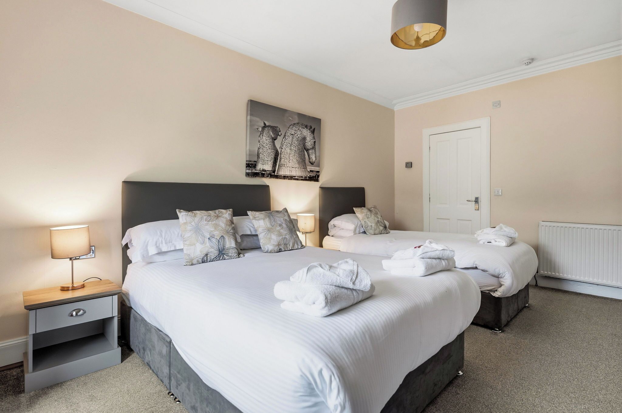 A hotel room with two beds, a nightstand with a lamp, a radiator, and a door.