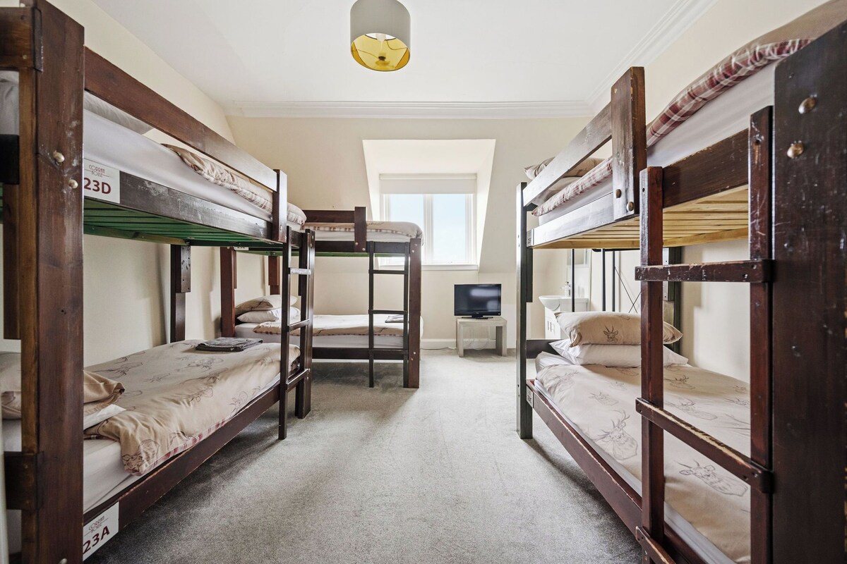 A dormitory room with bunk beds, a television on a small table, and a window providing natural light.