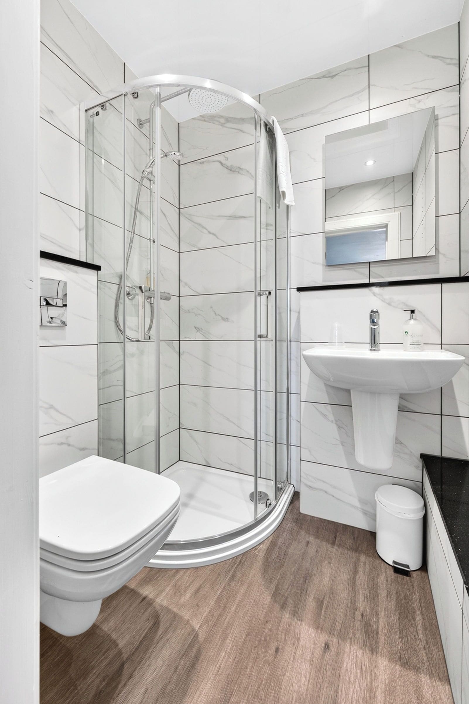 Un baño moderno con ducha envuelta en vidrio, inodoro blanco, lavamanos y espejo sobre el lavamanos.