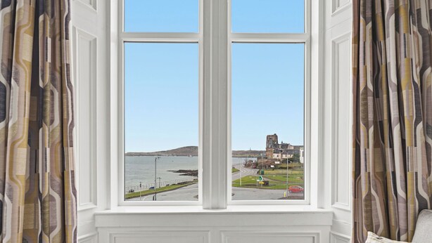 A room with a large window overlooking a coastal view, a chair, and patterned curtains. - Corran House Guest House (Oban)