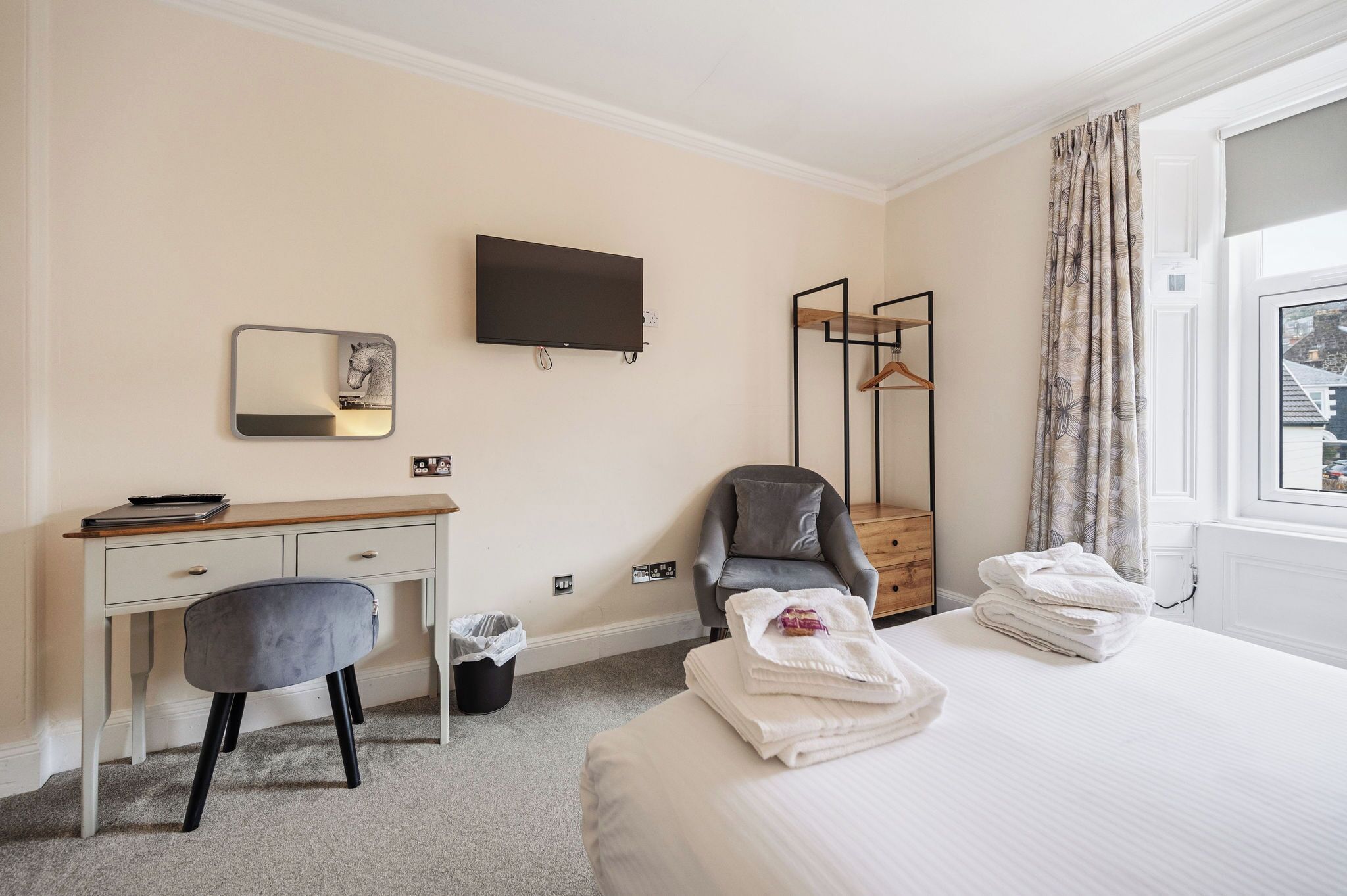 Habitación de hotel con cama, escritorio con silla, televisor, espejo, armario y ventana con cortinas.