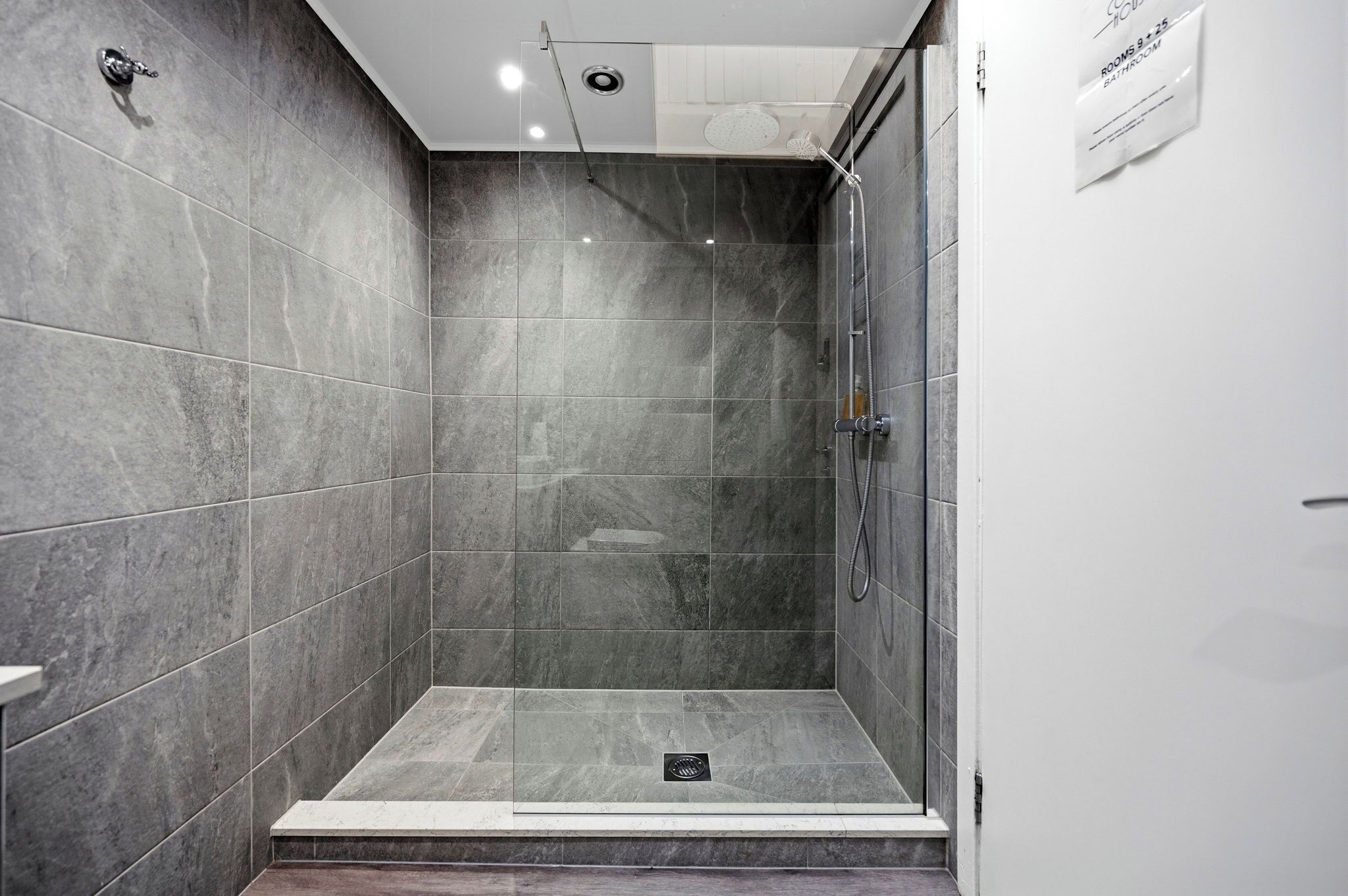A modern shower area with grey tiles, a glass door, and a sign indicating 'Rooms 3-5, 6-8, 9-11, 12-14, 15-17, 18-20, 21-23, 24-26, 27-29, 30-32, 33-35, 36-38, 39-41, 42-44, 45-47, 48-50, 51-53, 54-56, 57-59, 60-62, 63-65, 66-68, 69-71, 72-74, 75-77, 78-80, 81-83, 84-86, 87-89, 90-92, 93-95, 96-98, 99-101, 102-104, 105-107, 108-110