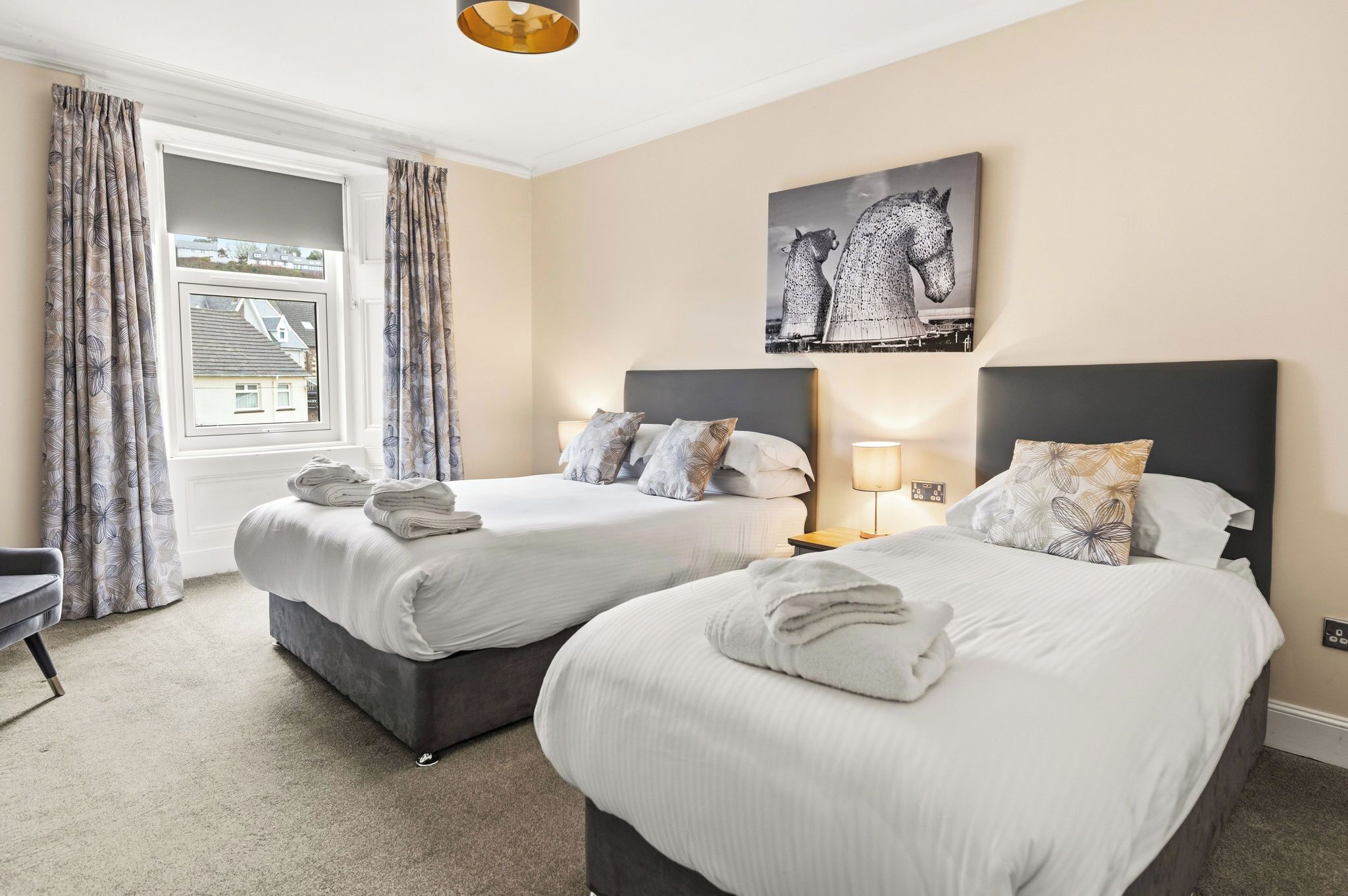 A hotel room with two beds, a window with curtains, a lamp, and a painting on the wall.