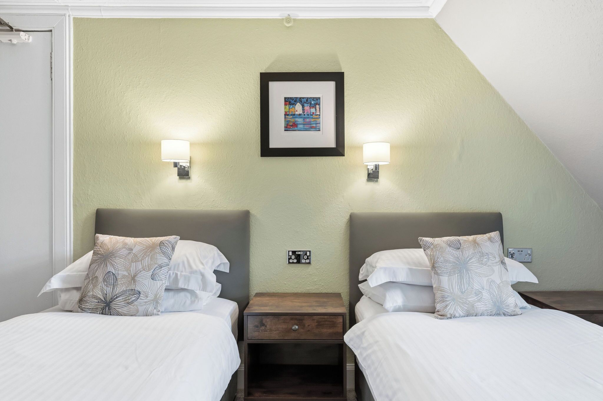 Une chambre d’hôtel avec deux lits, une table de chevet, un tableau encadré et des lampes fixées au mur.