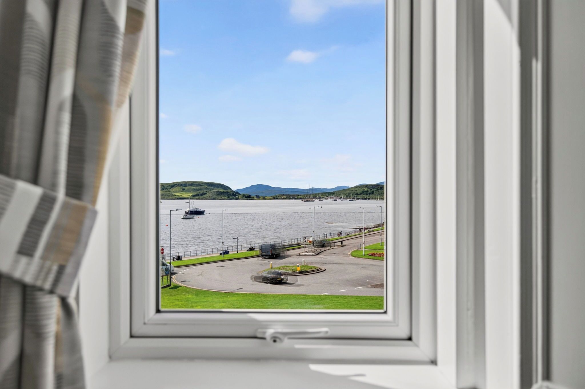 A view through a window of a coastal road with a body of water, a bridge, and distant hills.