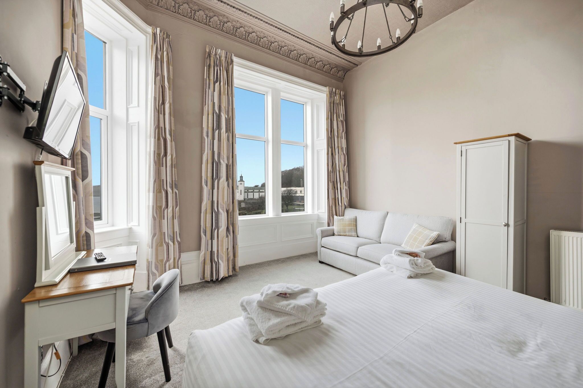 A hotel room with a bed, a desk with a computer, a chair, a wardrobe, and a view of the outside.