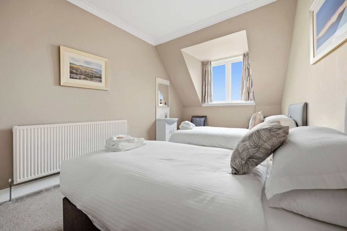 A bedroom with two beds, a radiator, a window with curtains, a framed picture on the wall, and a bedside table with a lamp.