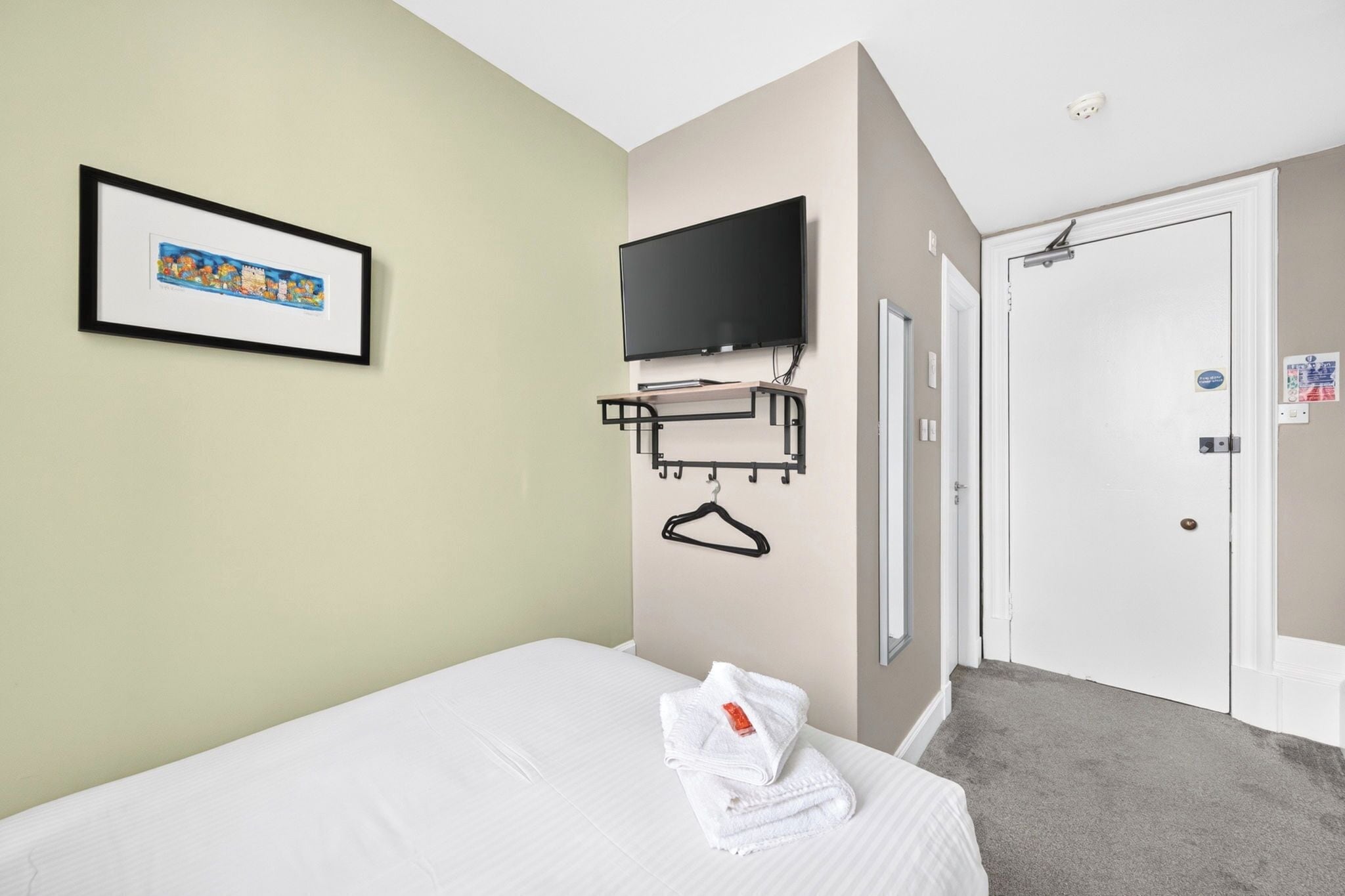 A hotel room with a bed, a mounted TV, a coat rack, and a door.