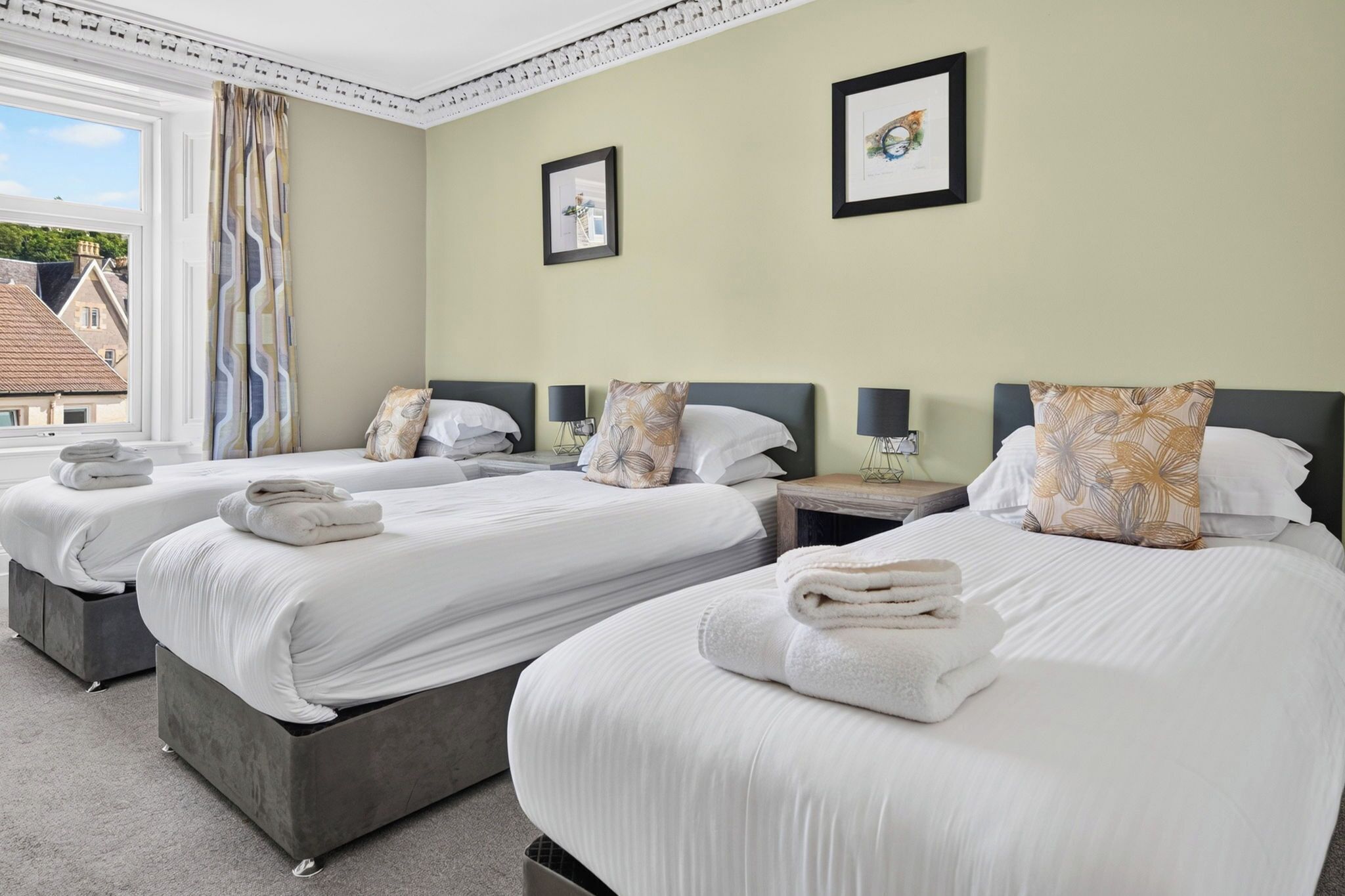 Een hotelkamer met drie bedden, elk voorzien van witte lakens en kussens, een raam met gordijnen en een lijst met een afbeelding aan de muur.
