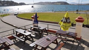 A waterfront dining area with picnic tables and umbrellas, overlooking a lake with boats and hills in the background. - Corran House Guest House (Oban)