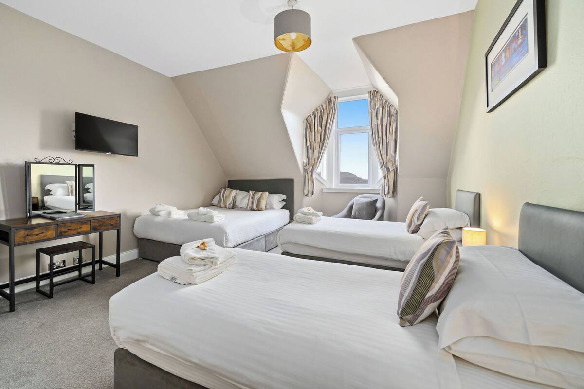 A hotel room with two beds, a desk with a computer, a TV mounted on the wall, and a window with curtains.