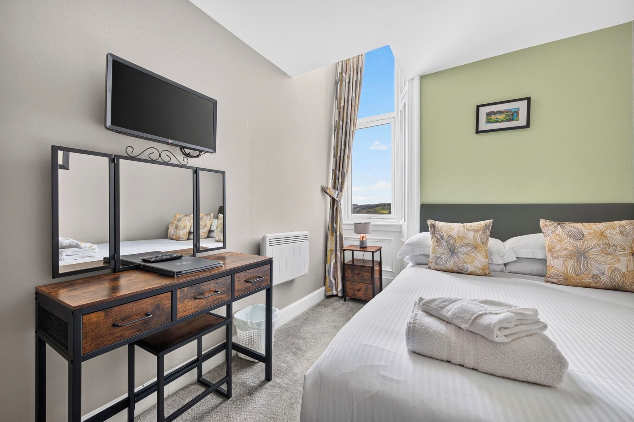 Une chambre d’hôtel avec un lit, un bureau avec un ordinateur, une télévision et une fenêtre avec des rideaux.