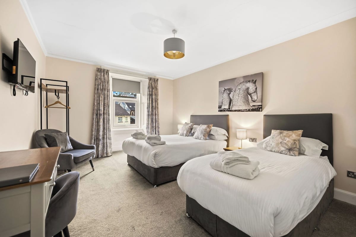 A hotel room with two beds, a desk, a chair, a TV, and a window with curtains.