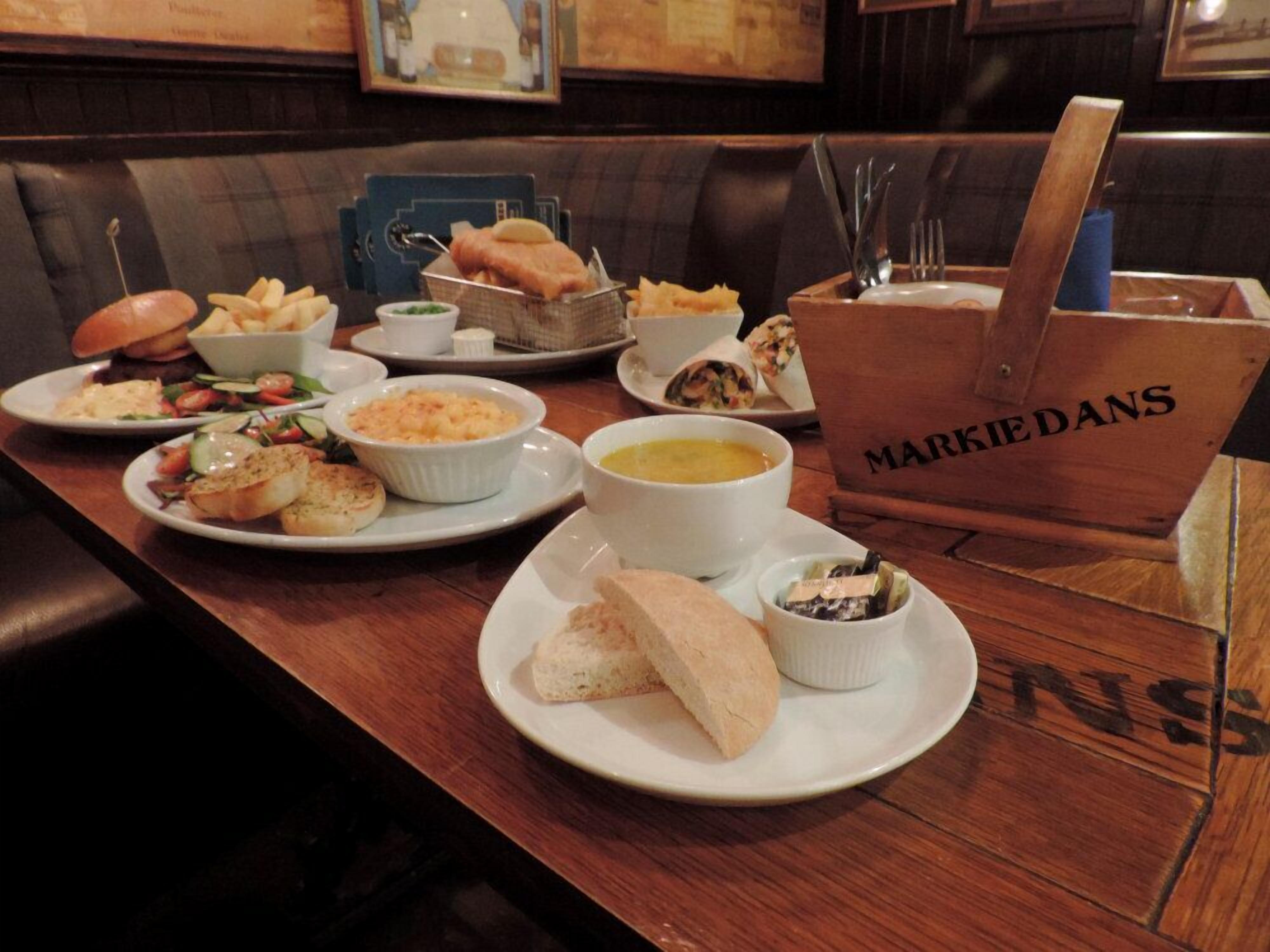 Un ambiente de restaurante con diversos platos en los platos, incluyendo pan, ensaladas y sopa. Hay un letrero con el nombre 'MARKIE DANS'.