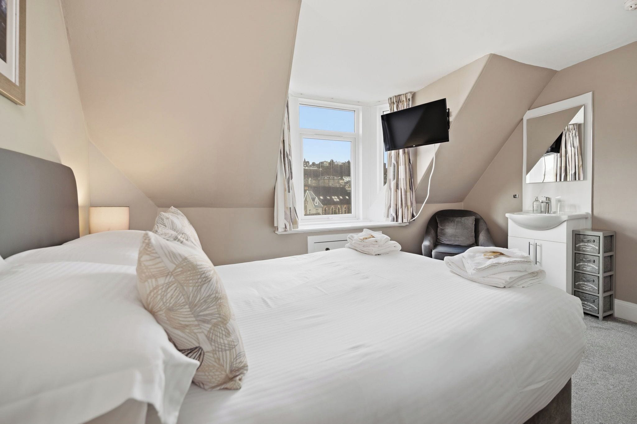A bedroom with a large bed, a TV mounted on the wall, a window with curtains, a chair, and a bedside table with a mirror.