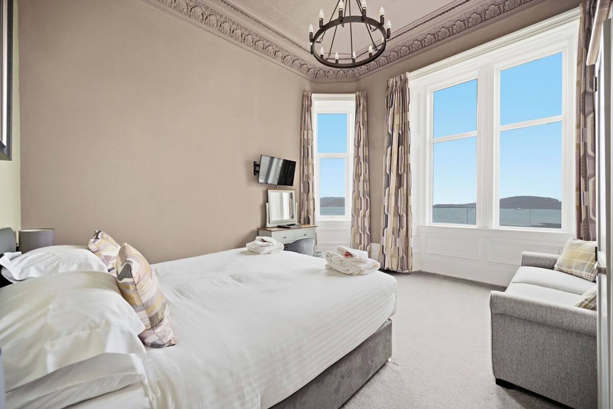 A bedroom with a large bed, a TV, a desk, and a view of the sea.