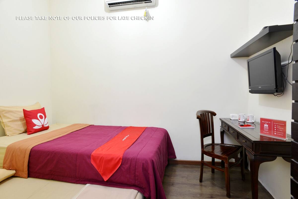 A hotel room with a bed, a desk, a chair, a television, and a sign with room amenities.