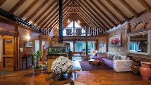 Une cabine spacieuse avec un plafond haut, des poutres en bois, un coin salon confortable avec un canapé et une table à manger, et une grande fenêtre qui laisse entrer la lumière naturelle.