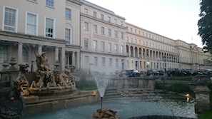 Ein klassischer Brunnen mit Statuen vor einem großen, mehrstöckigen Gebäude mit Rundbogenfenstern und Säulen.